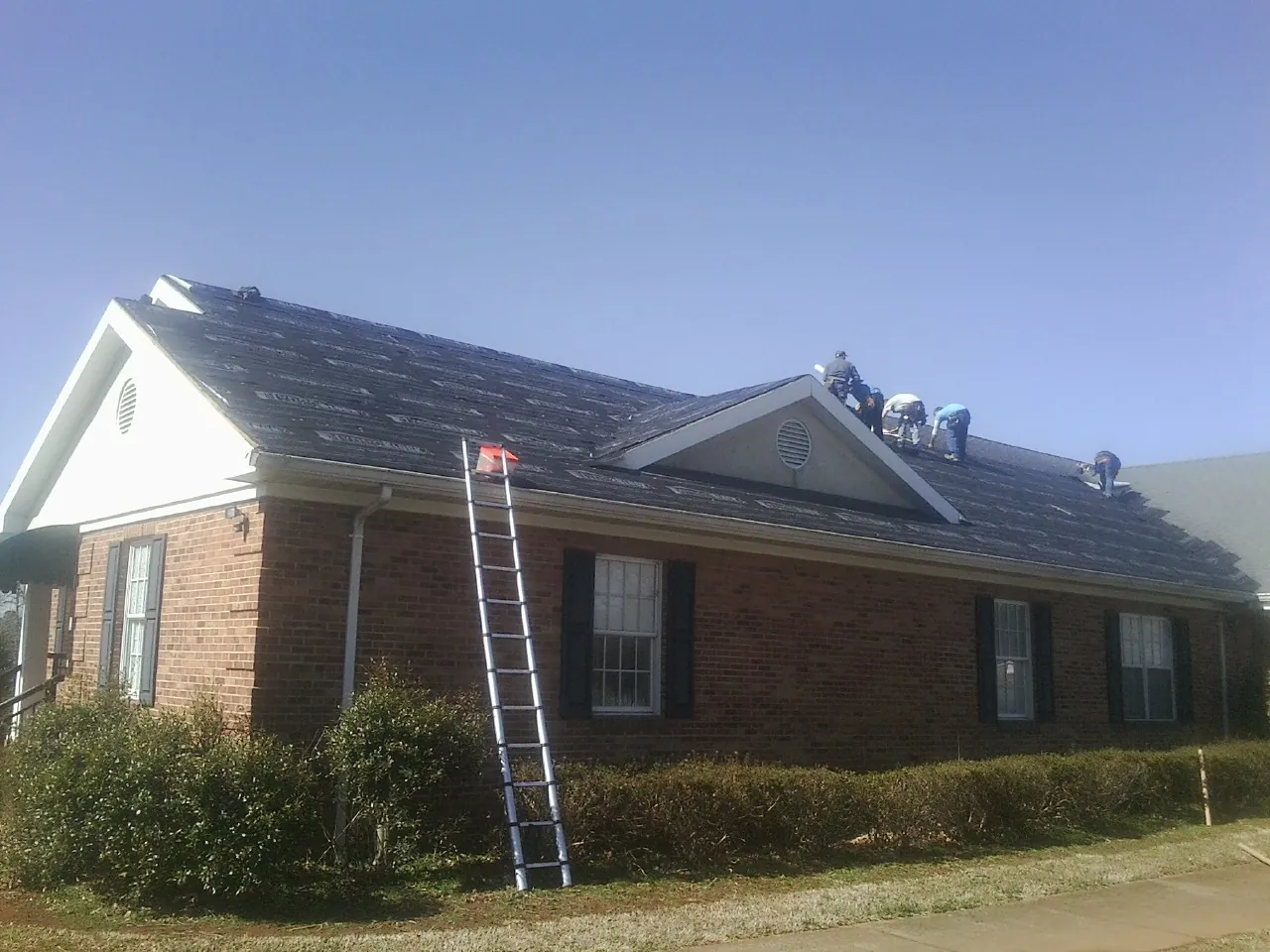 Crew tearing shingles off a brick ranch roof with a ladder set — Metal Roof Repair work by our crew in Sargent