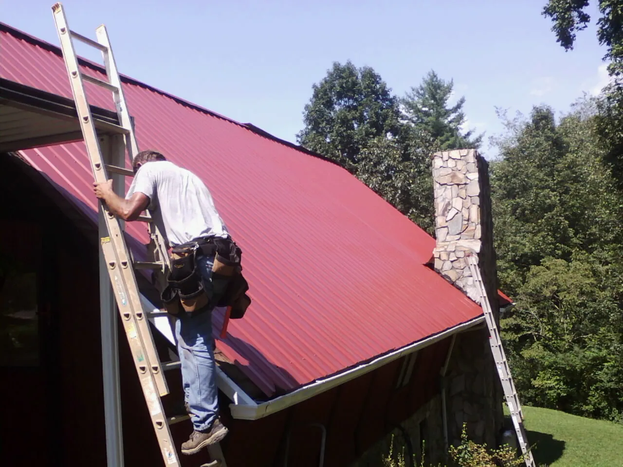 Roofer on a ladder beside a red metal roof and stone chimney — Soffit & Fascia Repair by Roofing Brothers in Sargent